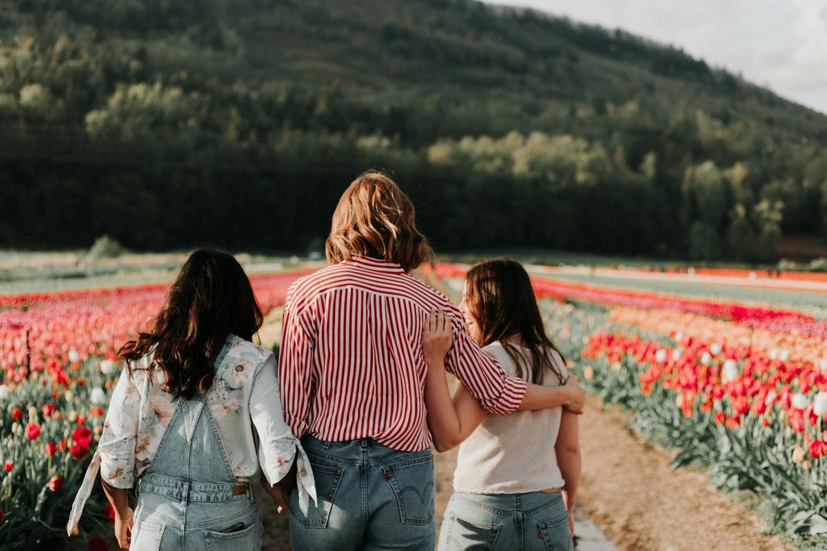 Three friends walking arm in arm along a path between rows of flowers in bloom