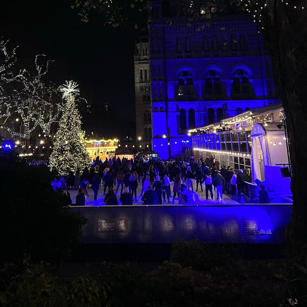 Skaters on a floodlit outdoor ice rink beside a lit tree at night